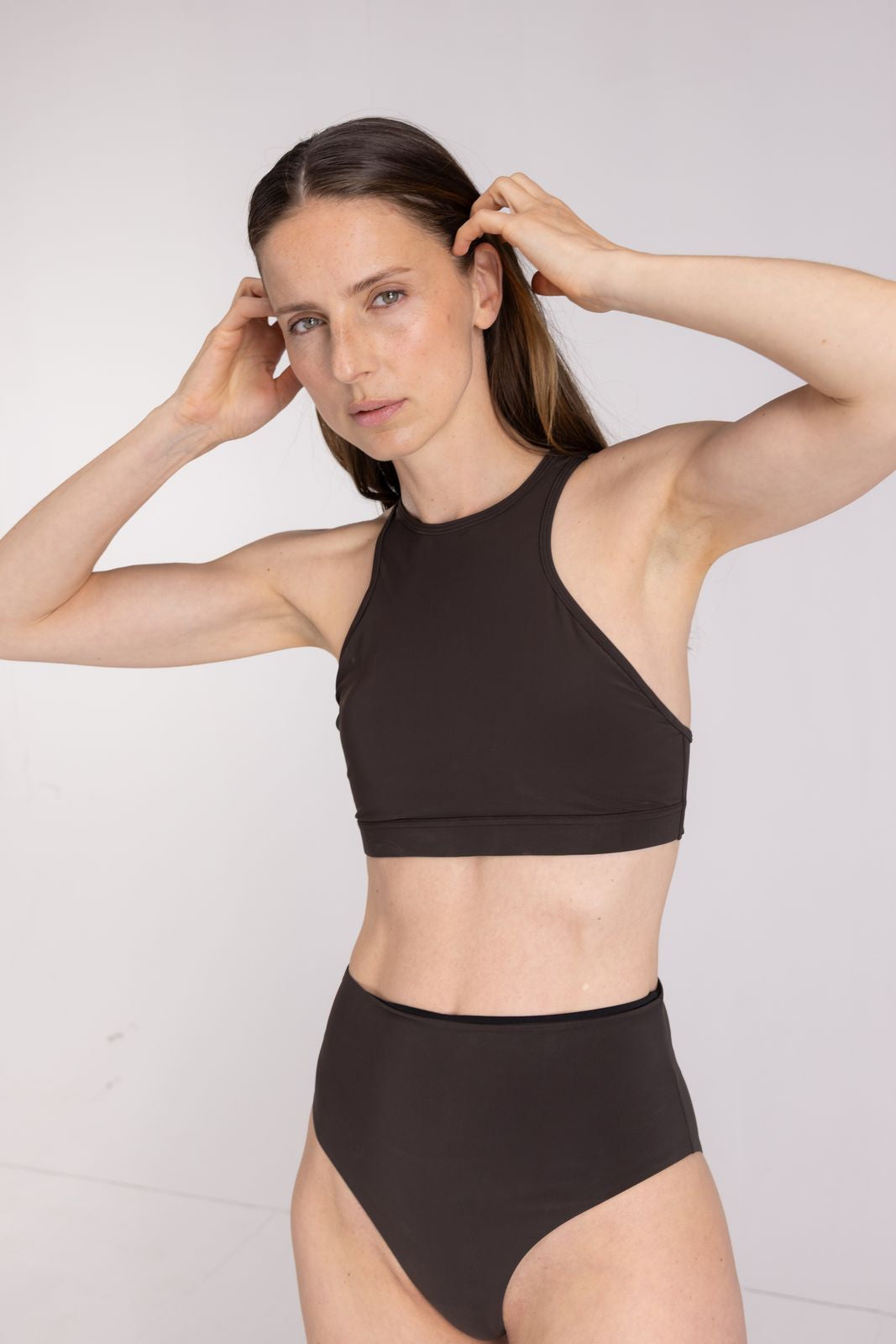 Woman in high-waisted coffee bikini bottom adjusting hair, sporty performance swim beachwear styling, on studio background