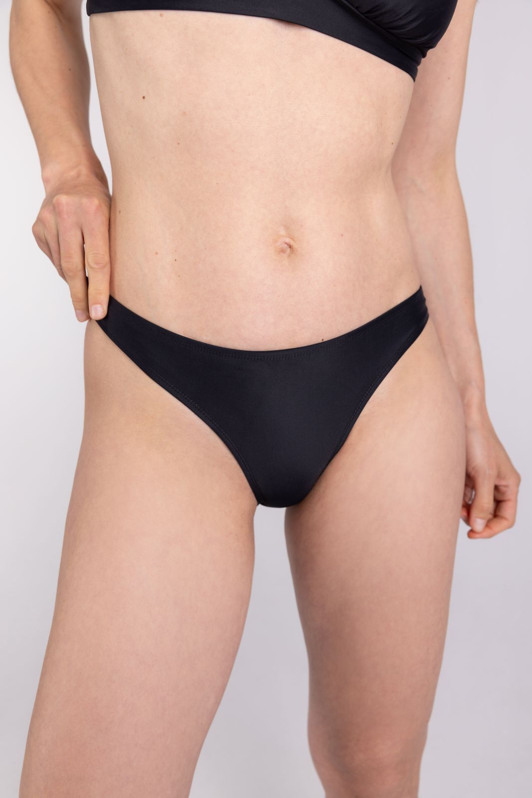 Woman wearing WALLIEN black bikini bottom in minimal studio setting, showing low waist fit and side profile, white backdrop
