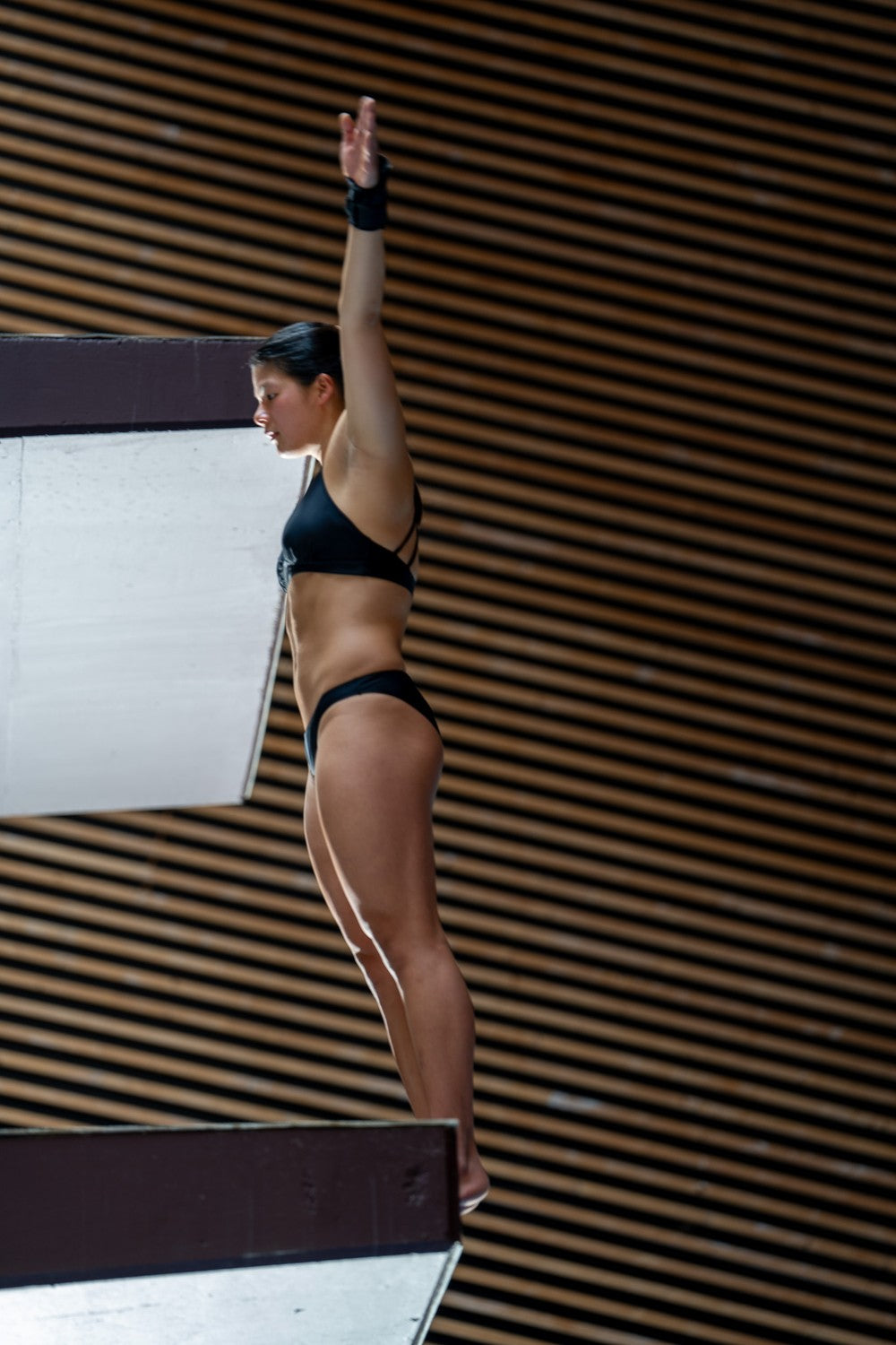 Woman in black bikini bottom raises arms on platform, preparing to dive with control and focus in indoor sports arena