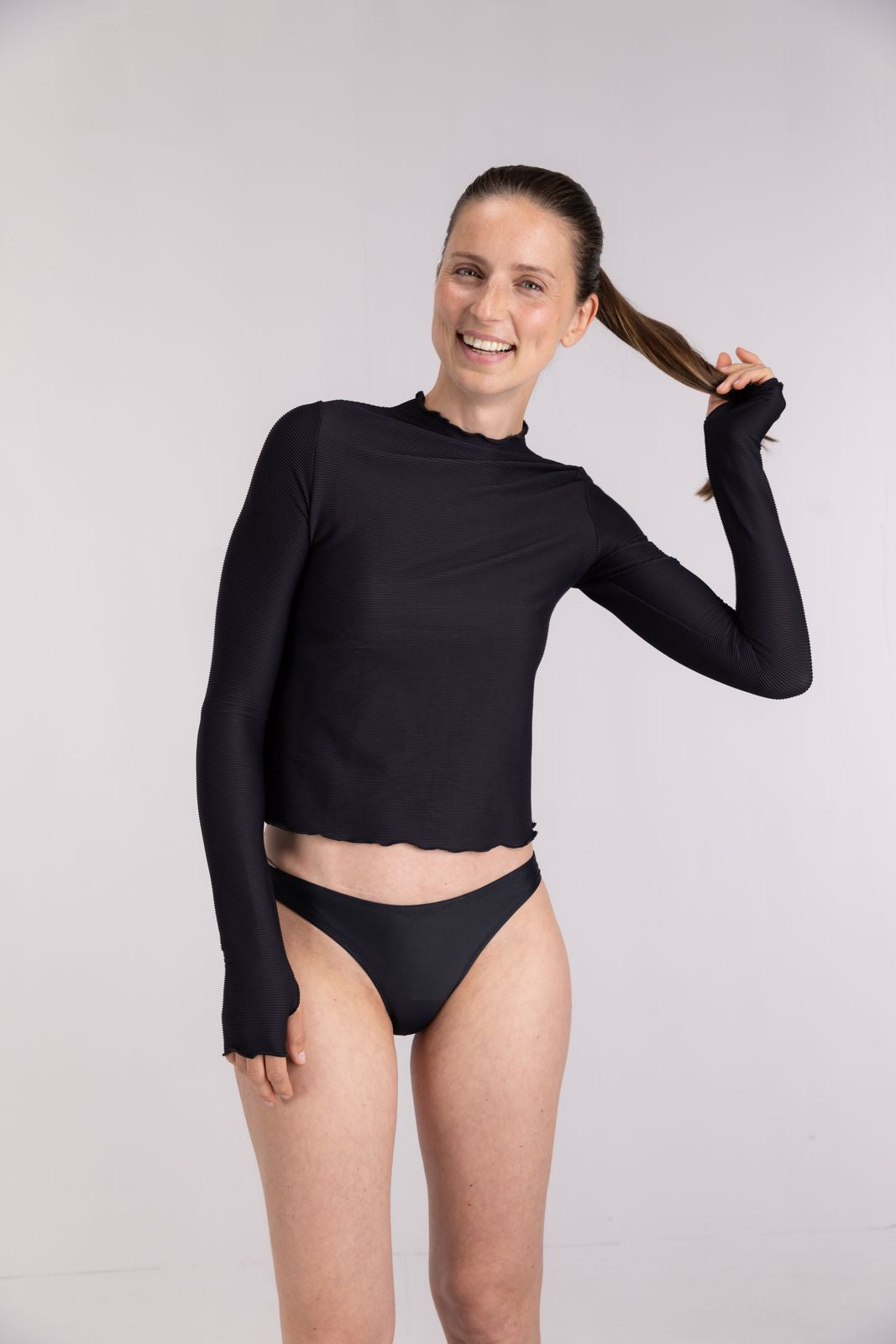 Smiling woman in WALLIEN black rib textured rash guard and bikini bottom, prepping for a surf session, in studio background
