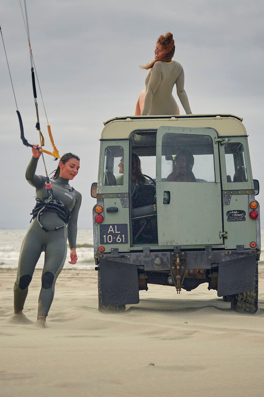 Women kitesurfing and resting on vintage car in Nikki van Dijk 3/2 wetsuit, with ocean and cloudy sky in background