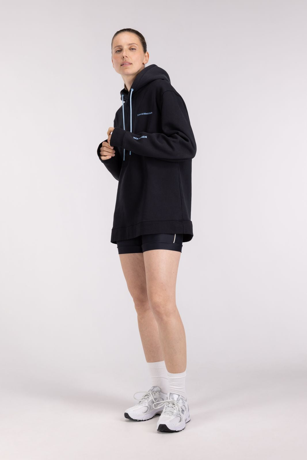 Woman in WALLIEN Black Ocean Hoodie warming up pre-adventure with blue drawstrings, standing indoors on studio background