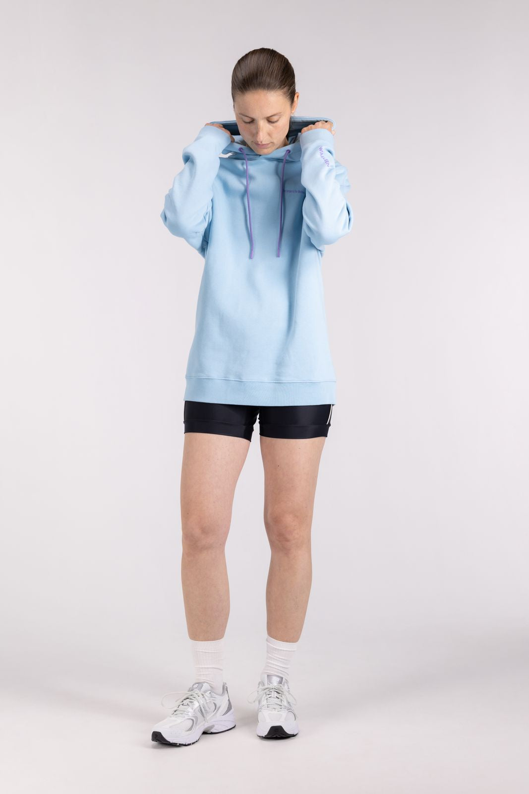 Model adjusting hood on WALLIEN Blue Ocean Hoodie, cozy surfwear for chilly days, shot in minimal indoor studio background