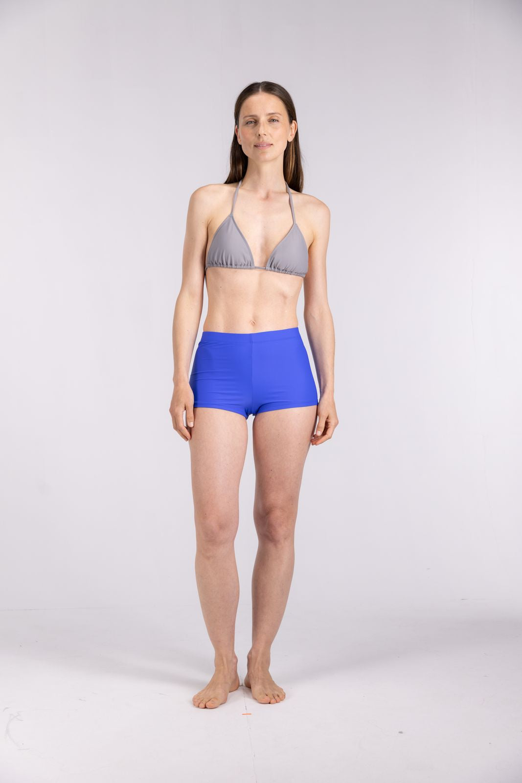 Woman modeling seal grey triangle bikini top and cobalt swim shorts, minimal eco-conscious fashion in studio background