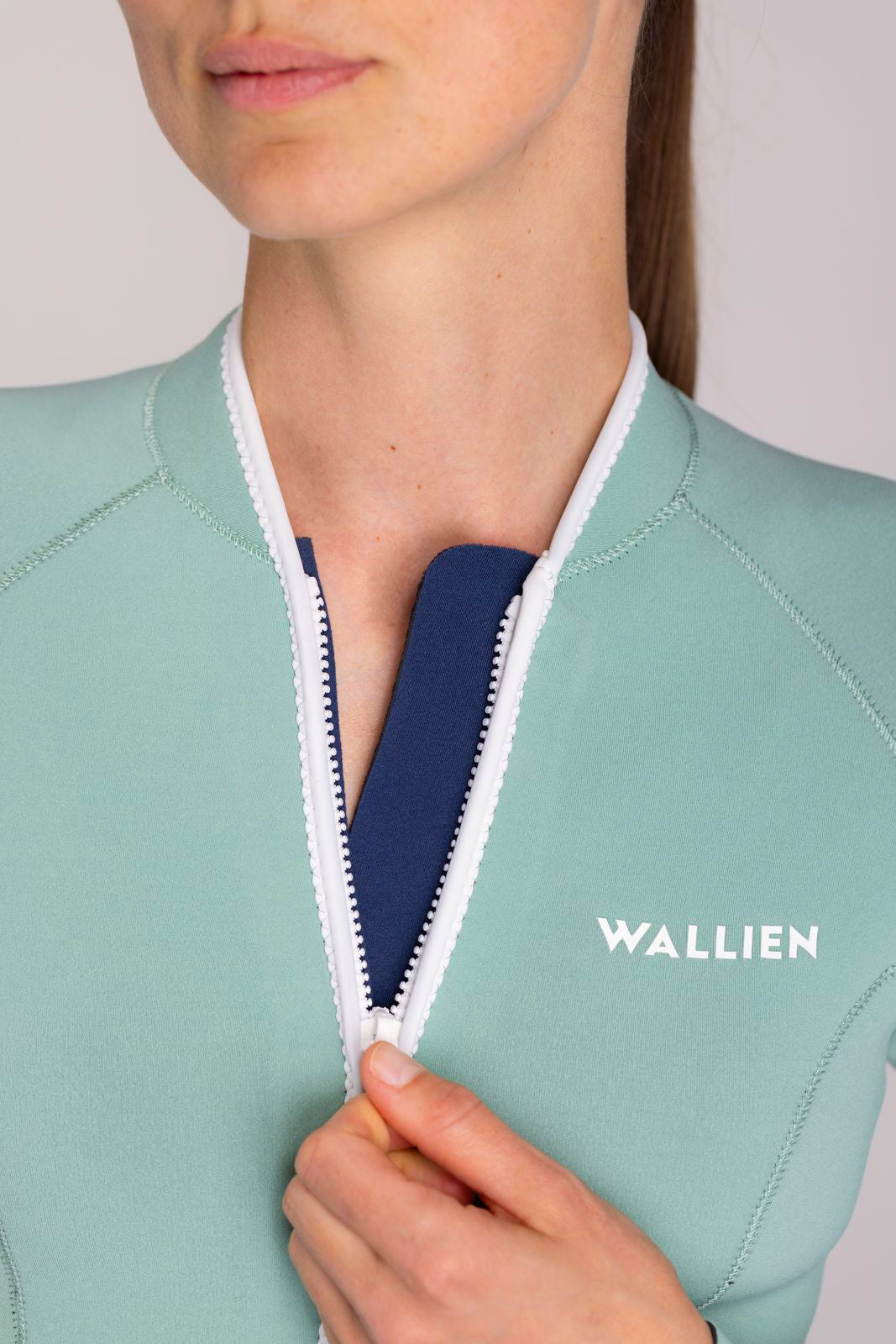 Close-up of aquamarine Yulex® springsuit wetsuit with white zip and WALLIEN logo, model holding zipper, on studio background