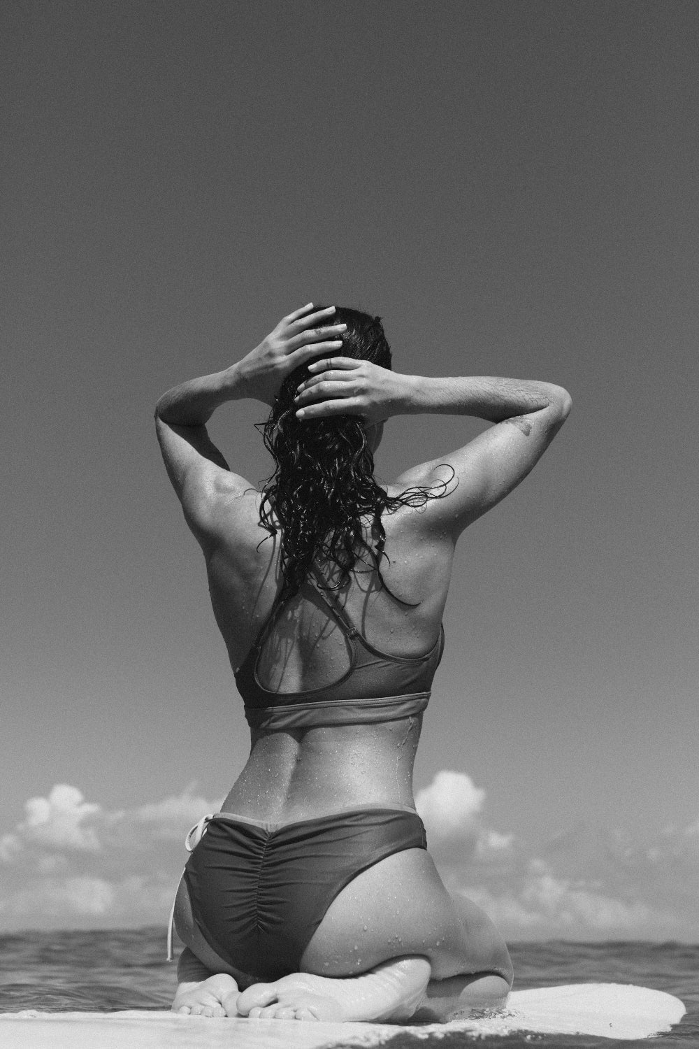 Back view of woman in cross-back bikini top sitting on surfboard, ocean horizon visible in grayscale background