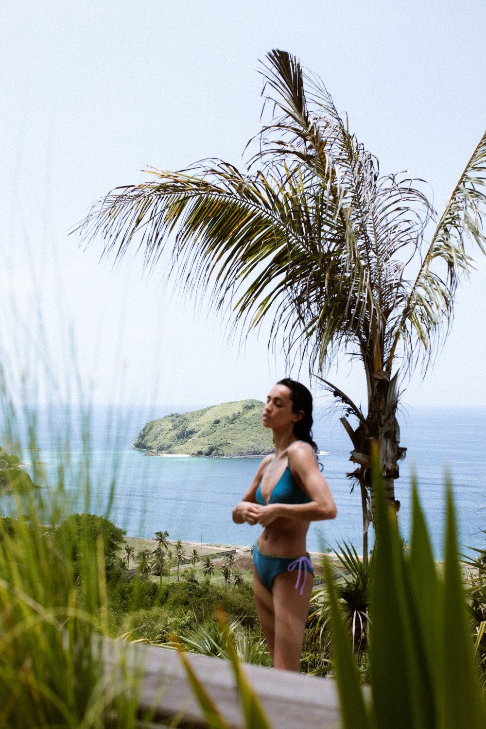 Woman in teal-green bikini top for ocean swimwear standing by palm trees, with tropical coastline and sea in background