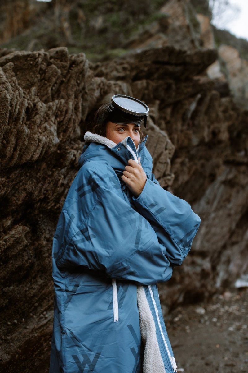 Woman in WALLIEN blue changing robe warming up post-swim, rugged coastal rocks in background after water activity