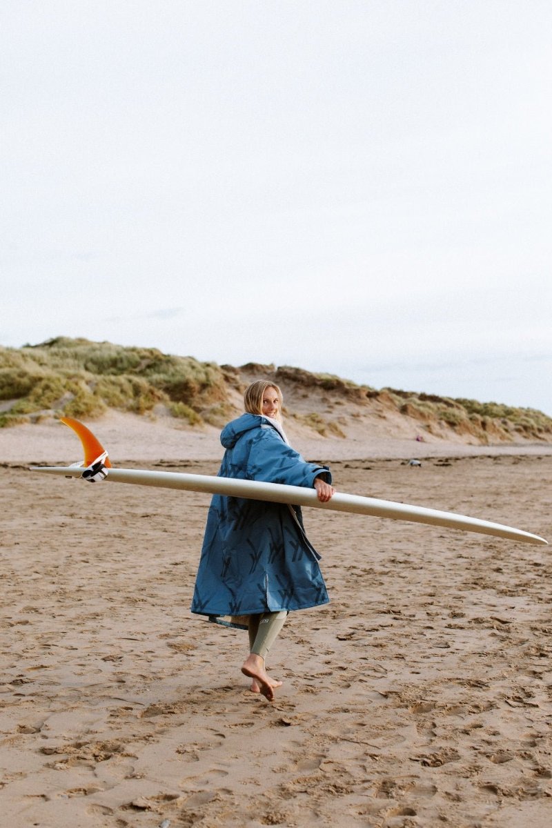 Woman carrying surfboard while wearing WALLIEN blue changing robe after session, walking barefoot on beach, with dunes behind