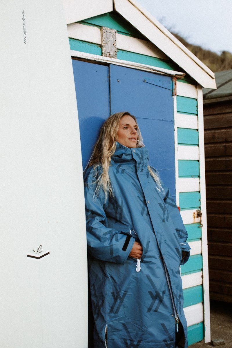 Woman in WALLIEN blue changing robe resting after swim, leaning on striped beach hut with surfboard in background