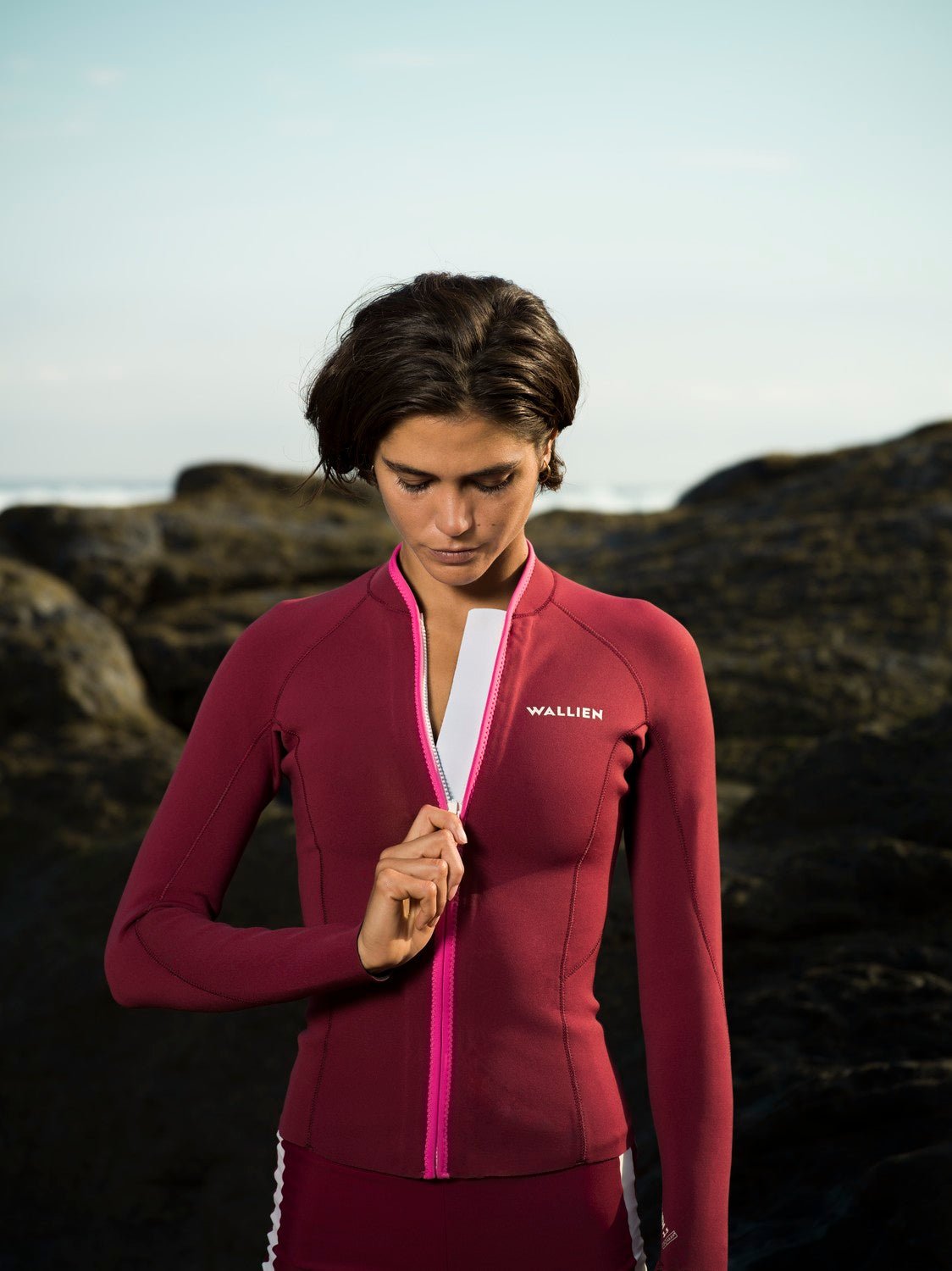 Woman zipping eco-friendly red Yulex® wetsuit jacket on coastal rocks at sunrise, ocean breeze in background