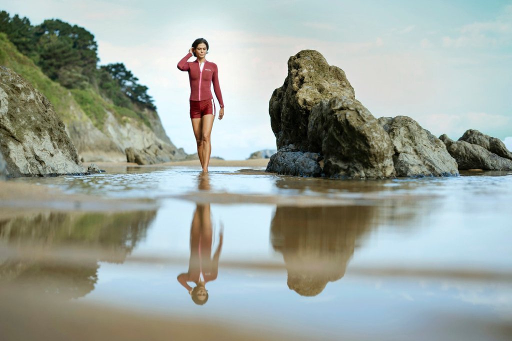 Woman walking barefoot in red Yulex® wetsuit jacket, beach cliffs and tidepool reflections around her