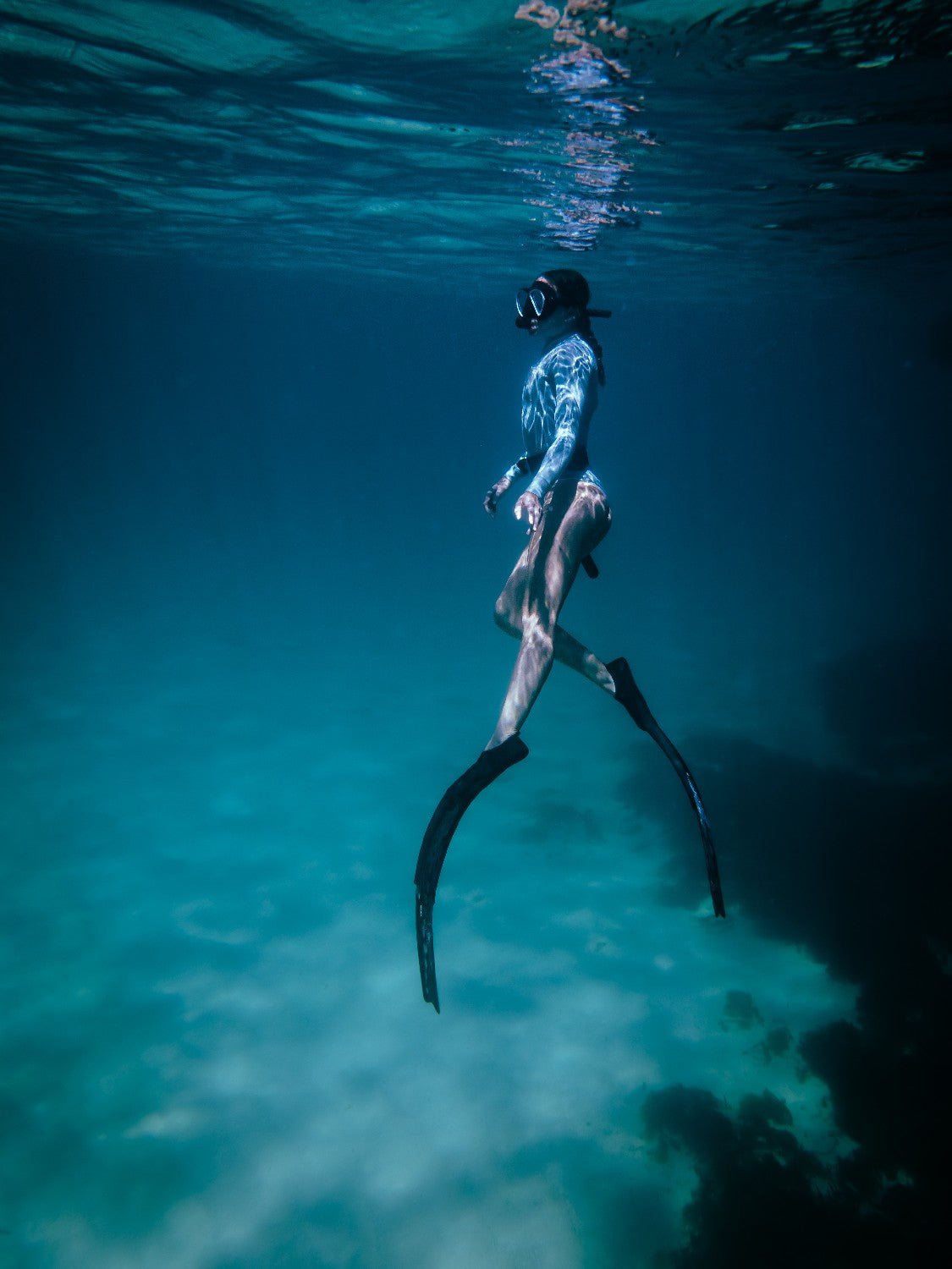 Freediver in aquamarine eco wetsuit with fins glides through open ocean, with coral reef and deep blue water in background