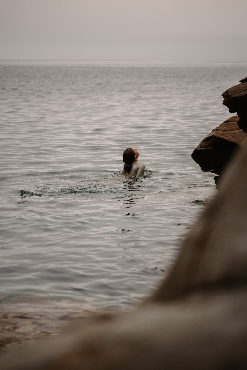 Woman swimming in open water wearing a beige-colored WALLIEN wetsuit with curved seams, stretch fit, rocky shoreline behind