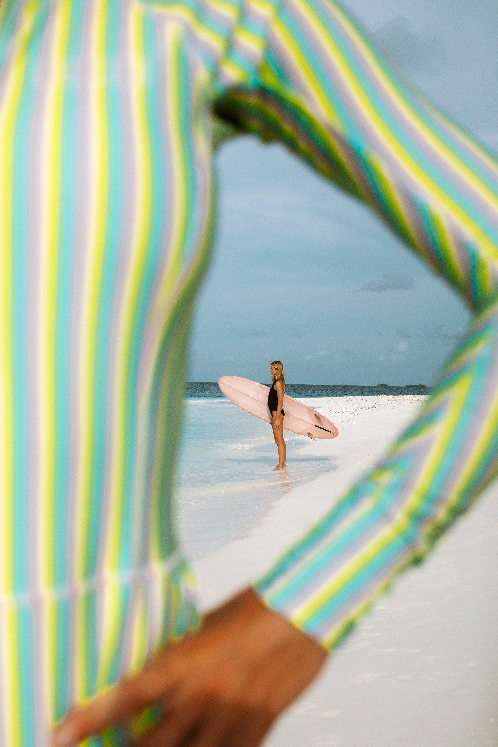 Surfer in a multi-color striped long sleeve WALLIEN swimsuit walking on white sand, framed by the surfer’s arm, ocean behind