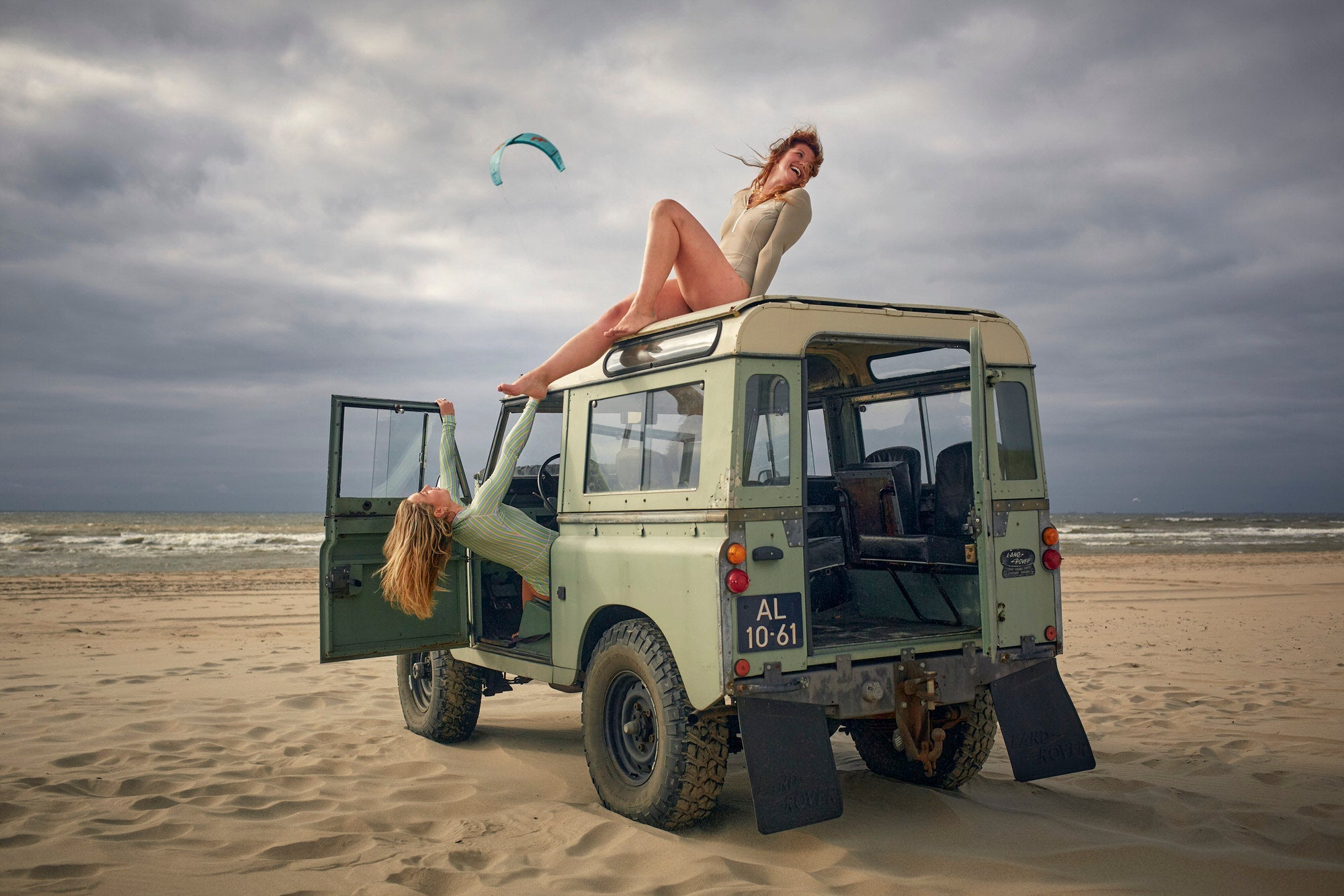Woman laughing in a colorful long sleeve WALLIEN swimsuit atop a Land Rover, with a kite and North Sea beach in background