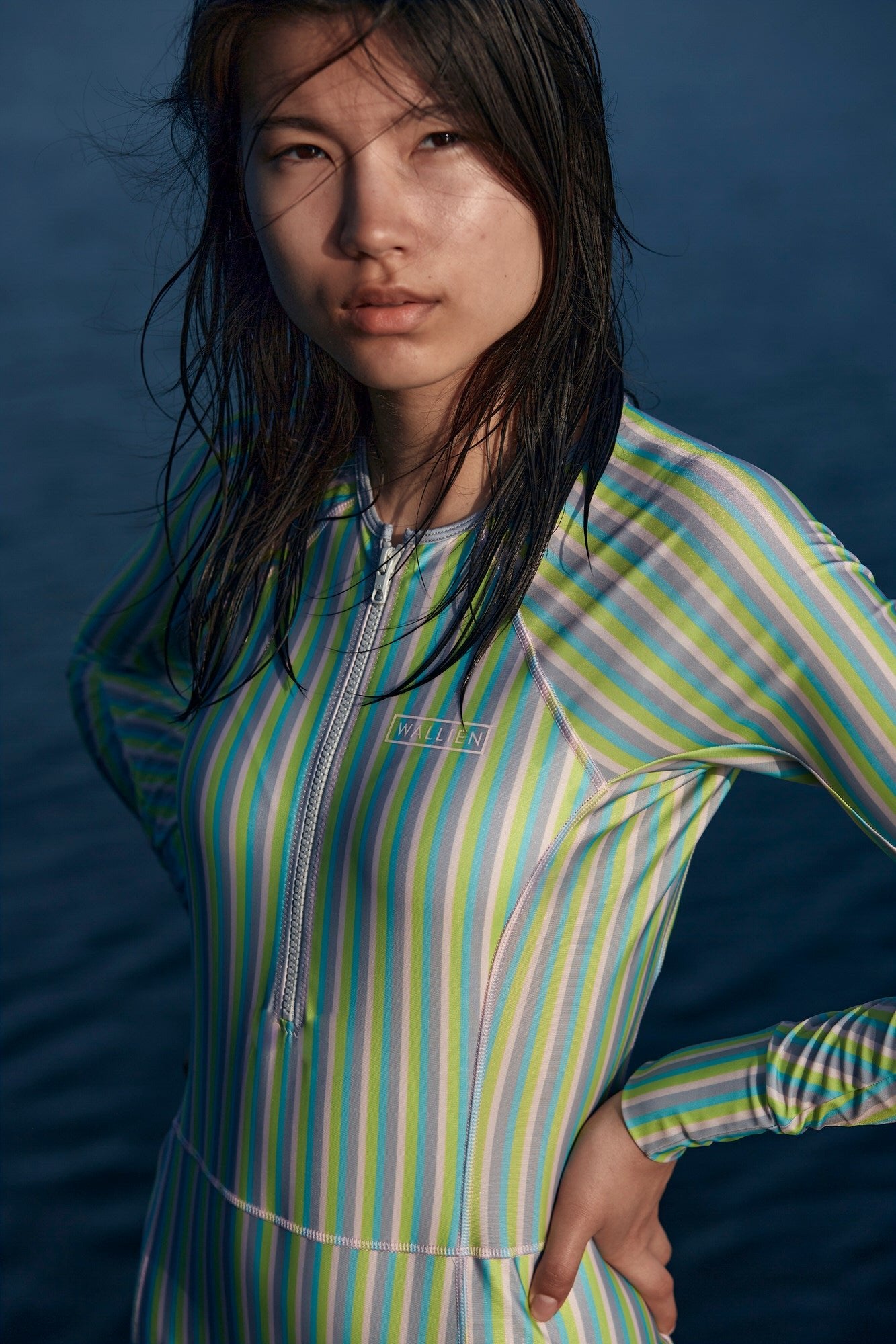 Woman wearing WALLIEN striped long swimsuit looking forward, with wet hair and calm lake water softly blurred in background