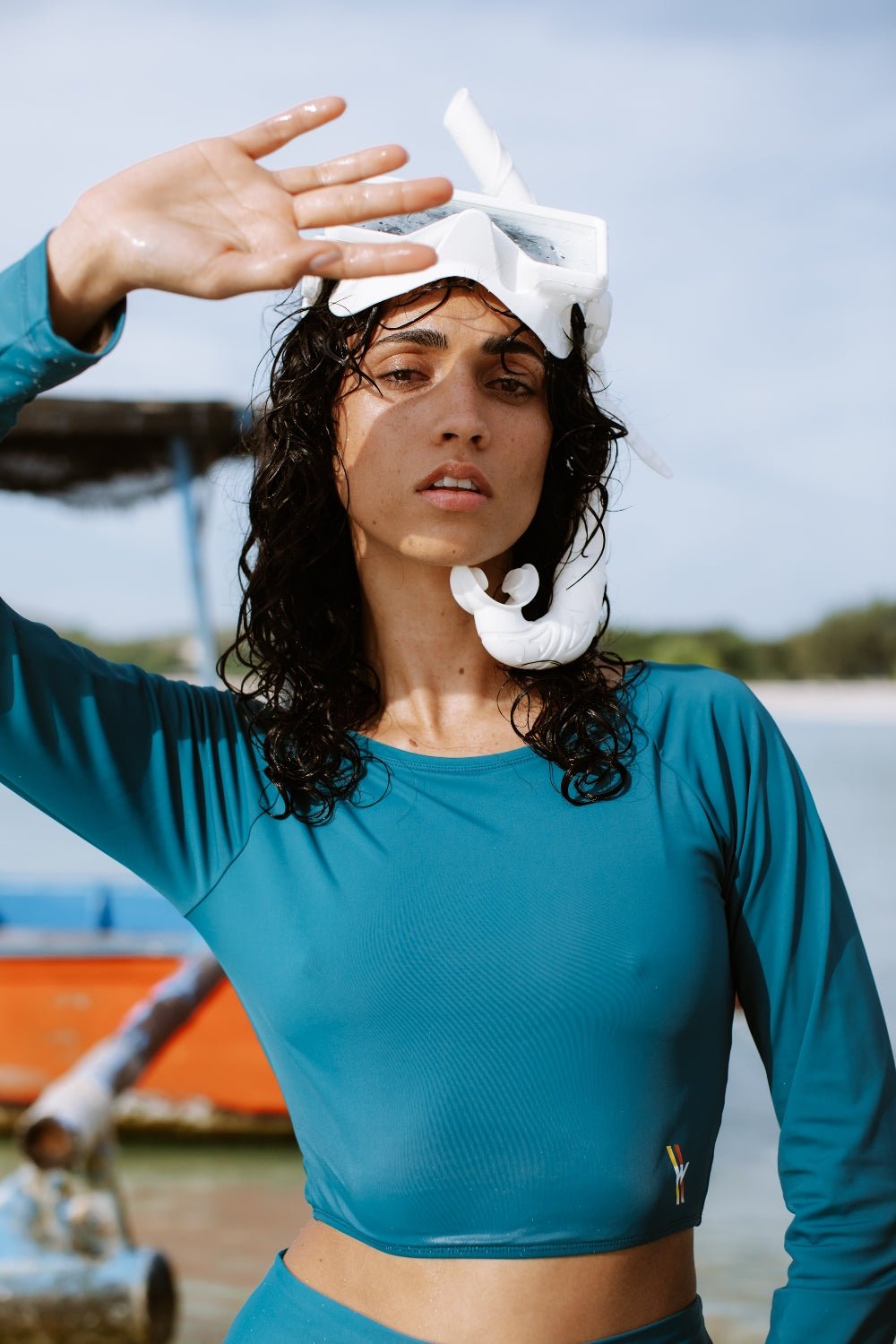 Woman in teal WALLIEN rash guard top adjusting snorkel mask by the beach, sunlit ocean with boats and sky in background