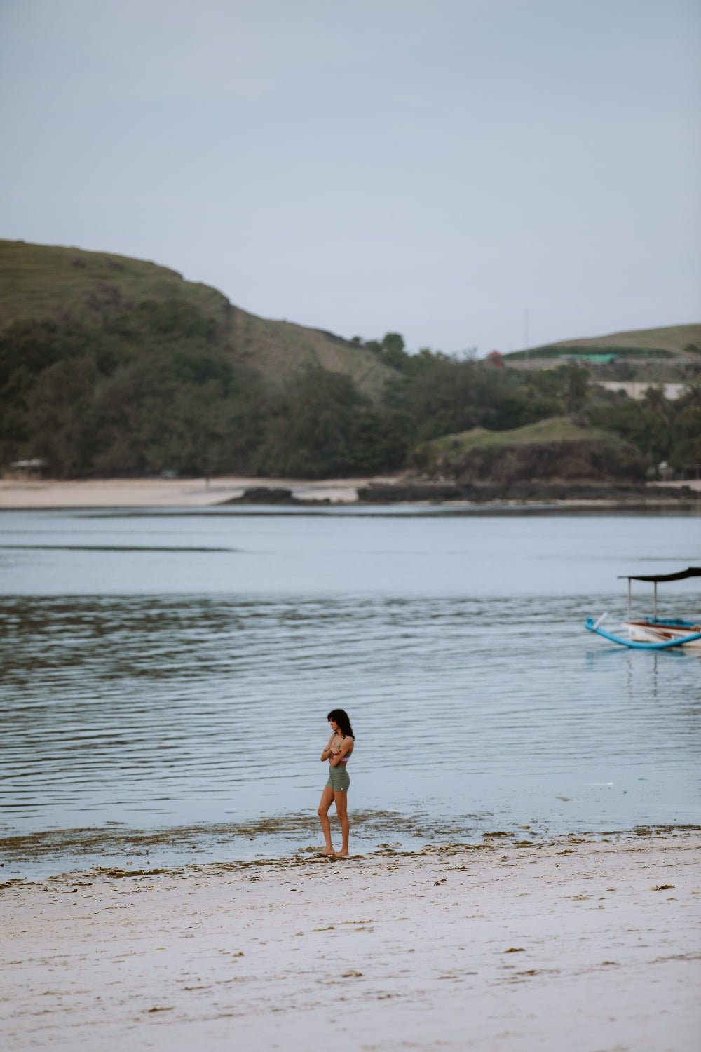 Woman walking along wet sand in green performance sport shorts, with lush hills and calm sea in the background