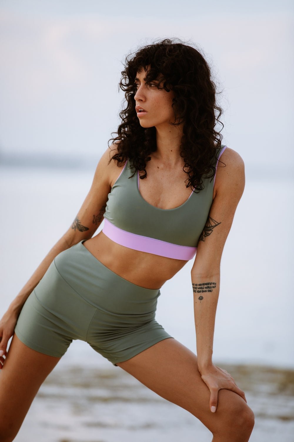 Woman stretching in green eco-friendly sport shorts by the ocean, designed for water sports, cloudy sea in background