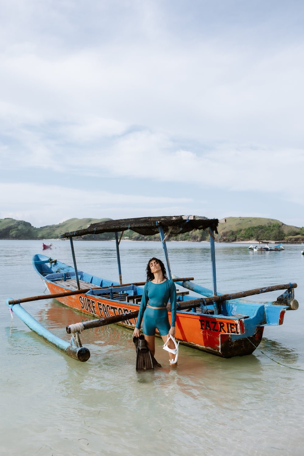 Woman in teal sport shorts and rashguard near surf boat on shore, bright day with island hills and sea in background