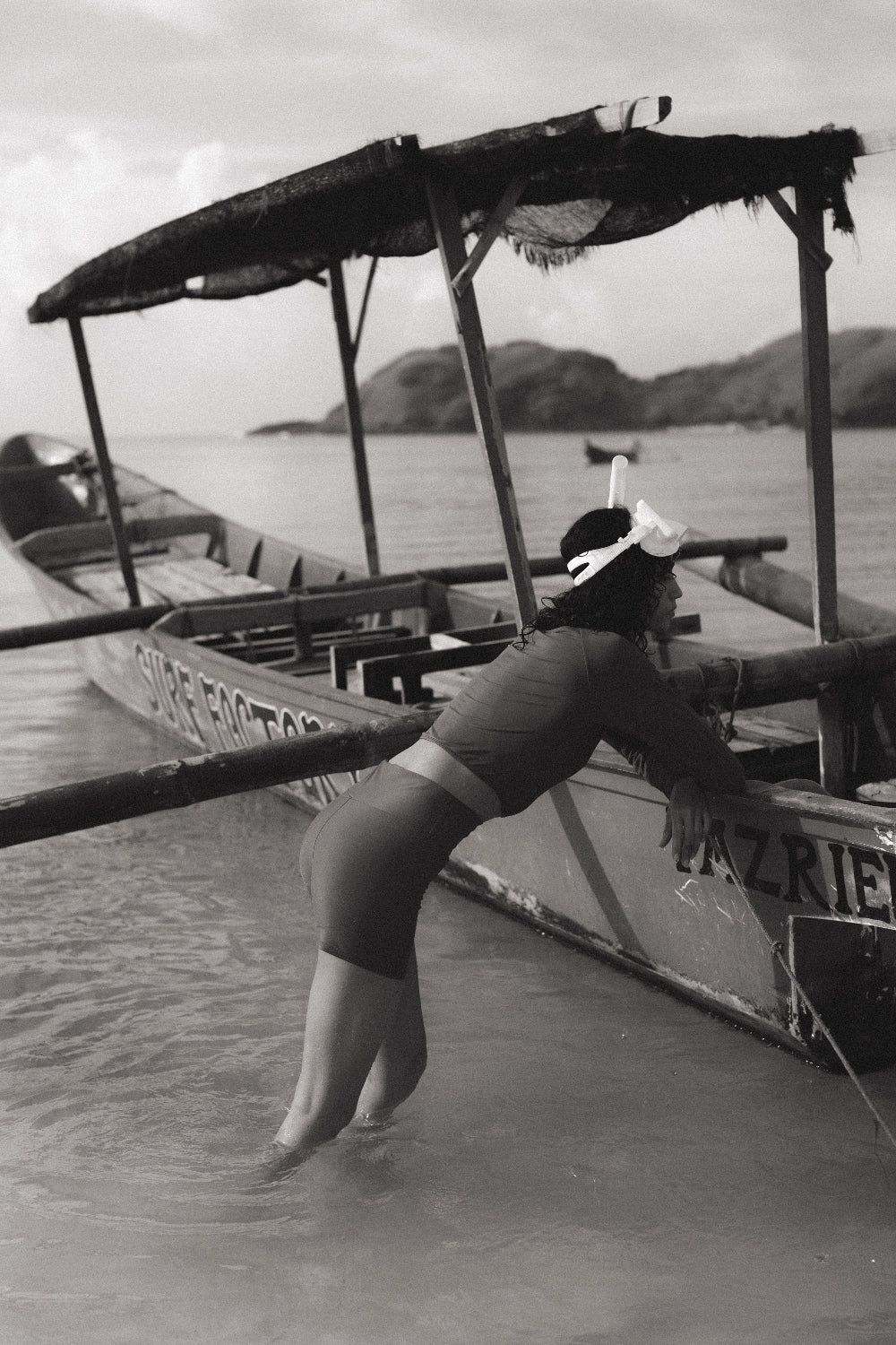 Teal sport shorts worn during beach break, woman leaning on surf boat in shallow water, island coastline in background