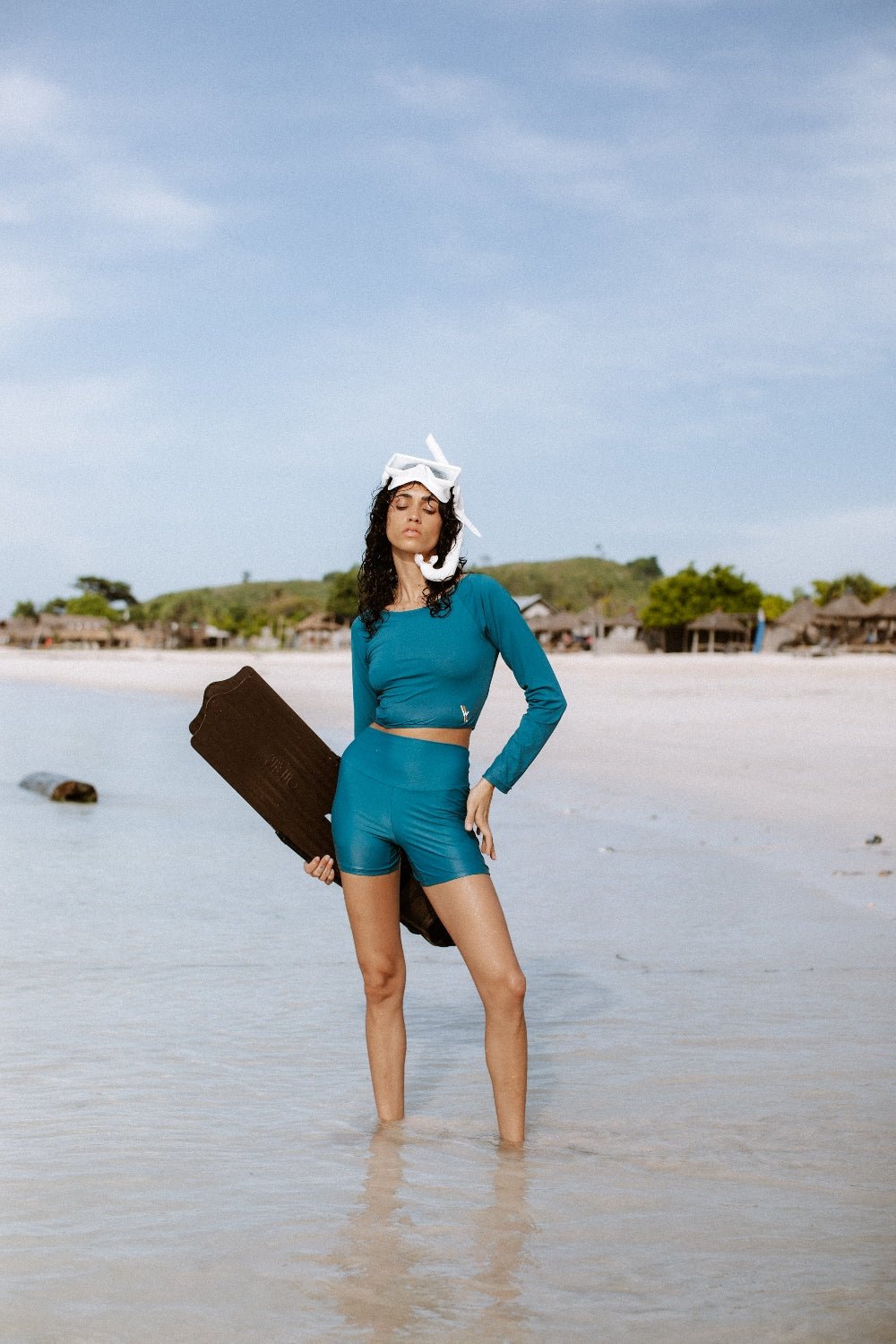 Woman in teal WALLIEN sport shorts holding fins on a sunny beach, ready for ocean swim, huts and sea in background