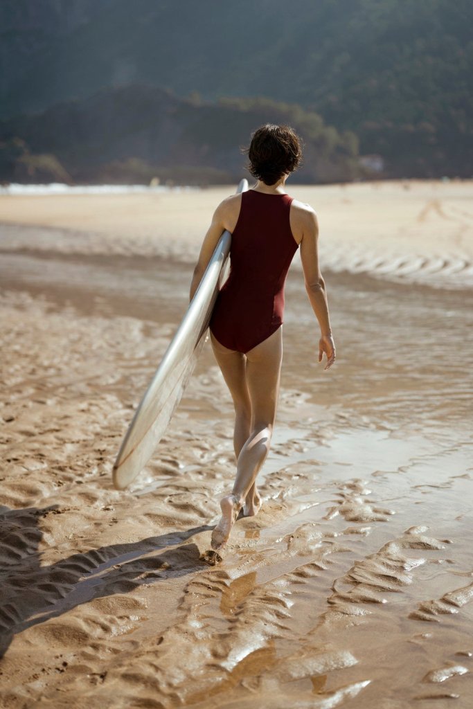 Back view of woman in red one-piece walking on beach with surfboard, combining function and coverage in eco swimwear style